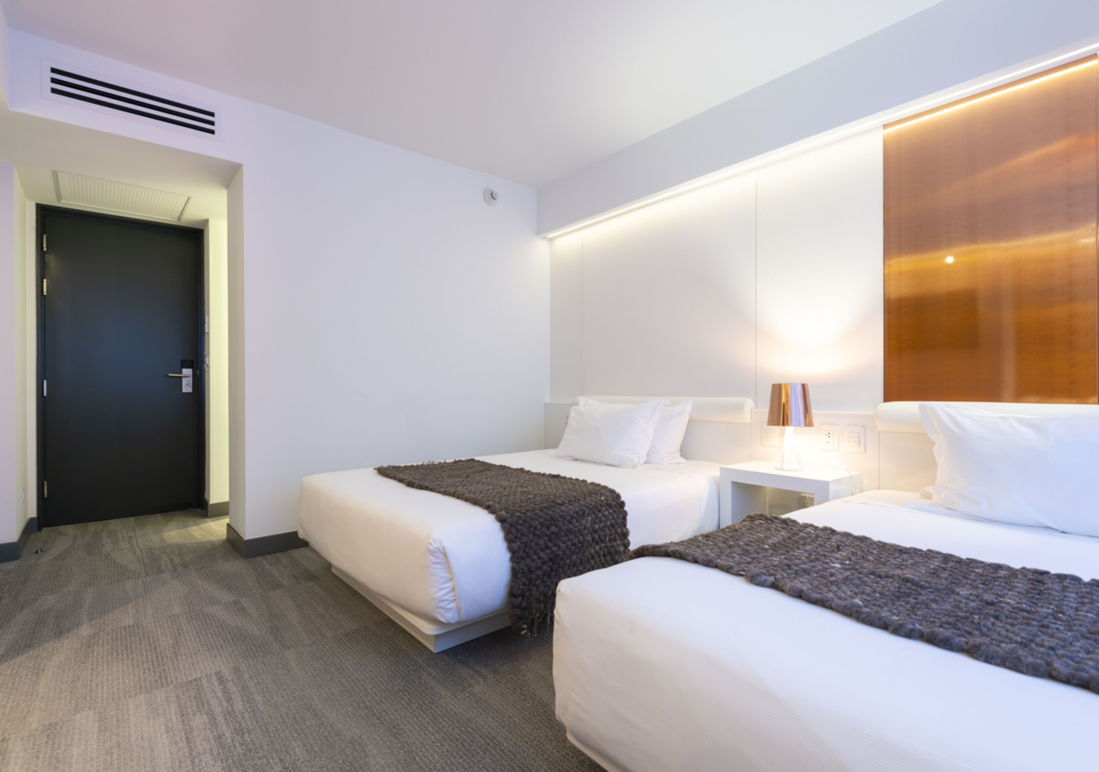 Ein Hotelzimmer mit zwei Betten, einer dunklen Tür, einer weißen Wand, einer Lampe und einem Teppichboden.
