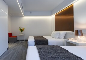 Una habitación de hotel moderna con dos camas, una mesita con lámpara, una silla roja y un florero con flores.