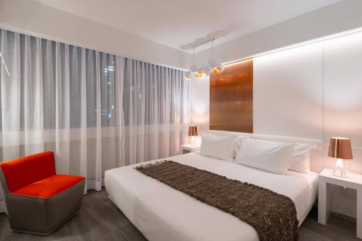 A modern hotel room with a large bed, a red chair, and a city view through sheer curtains.