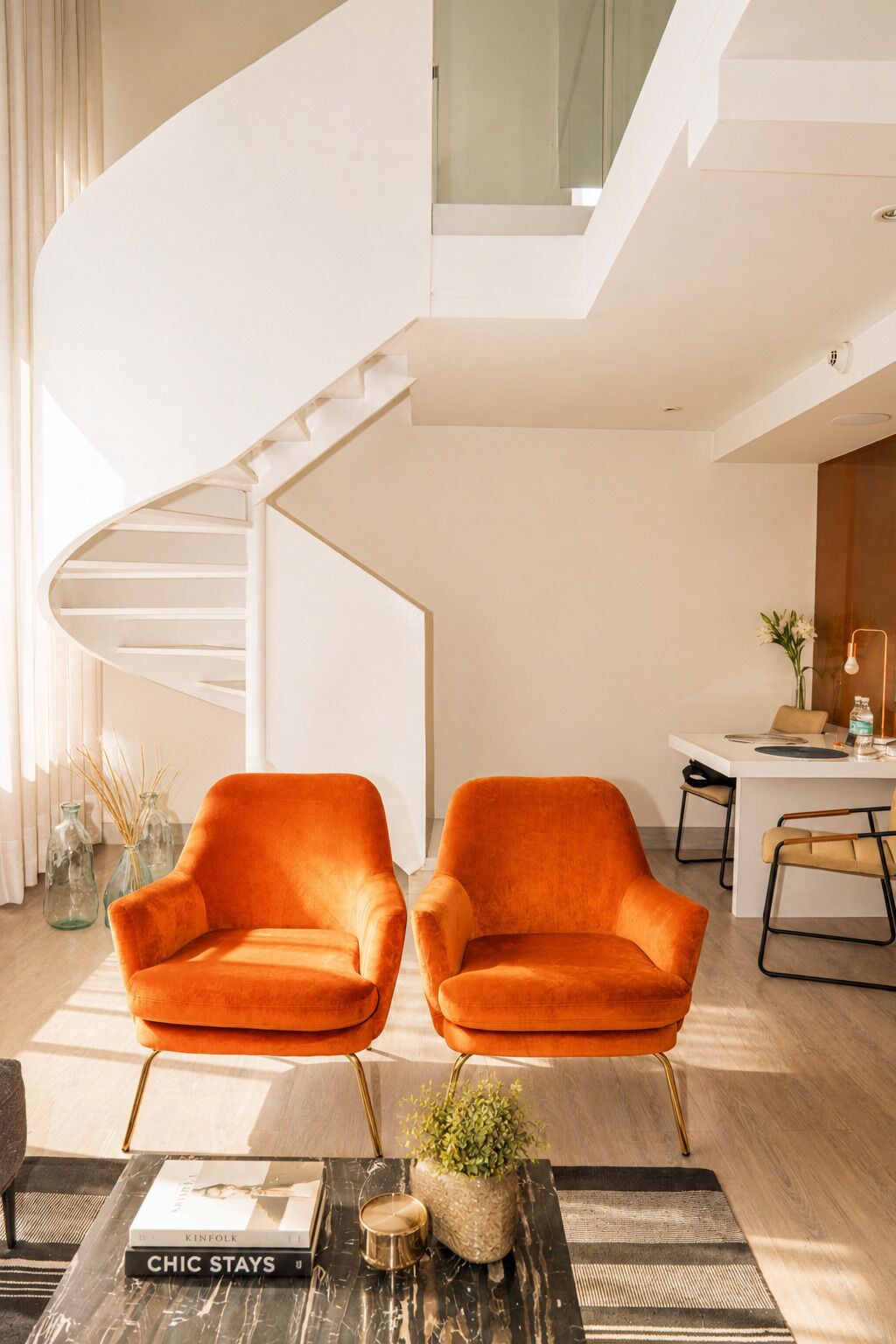 A modern interior with a spiral staircase, two orange armchairs, a dining area, and a coffee table with books and a candle.