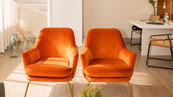 A modern interior with a spiral staircase, two orange armchairs, a dining area, and a coffee table with books and a candle.