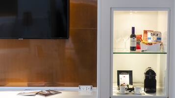 Zona de recepción de un hotel con un televisor montado en la pared, una vitrina de cristal que contiene una botella de vino, una cafetera y algunas revistas.