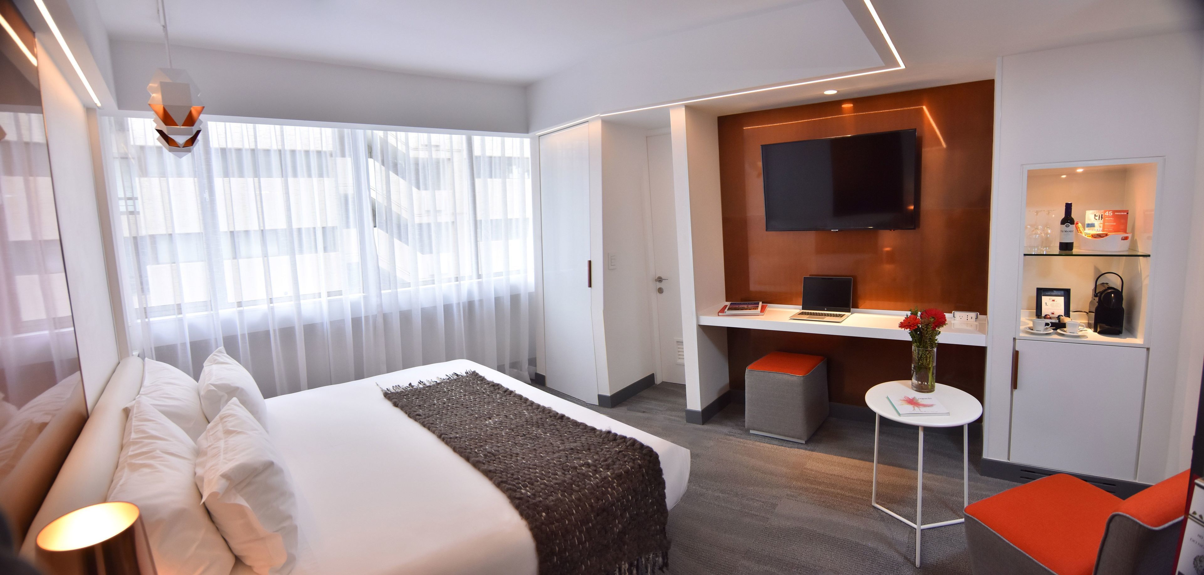 A modern hotel room with a bed, a desk, a TV, and a small kitchenette.