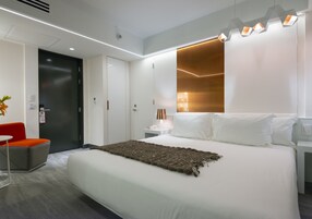 Una habitación de hotel moderna con una cama grande, una mesita de noche con lámpara, una silla roja y una puerta.
