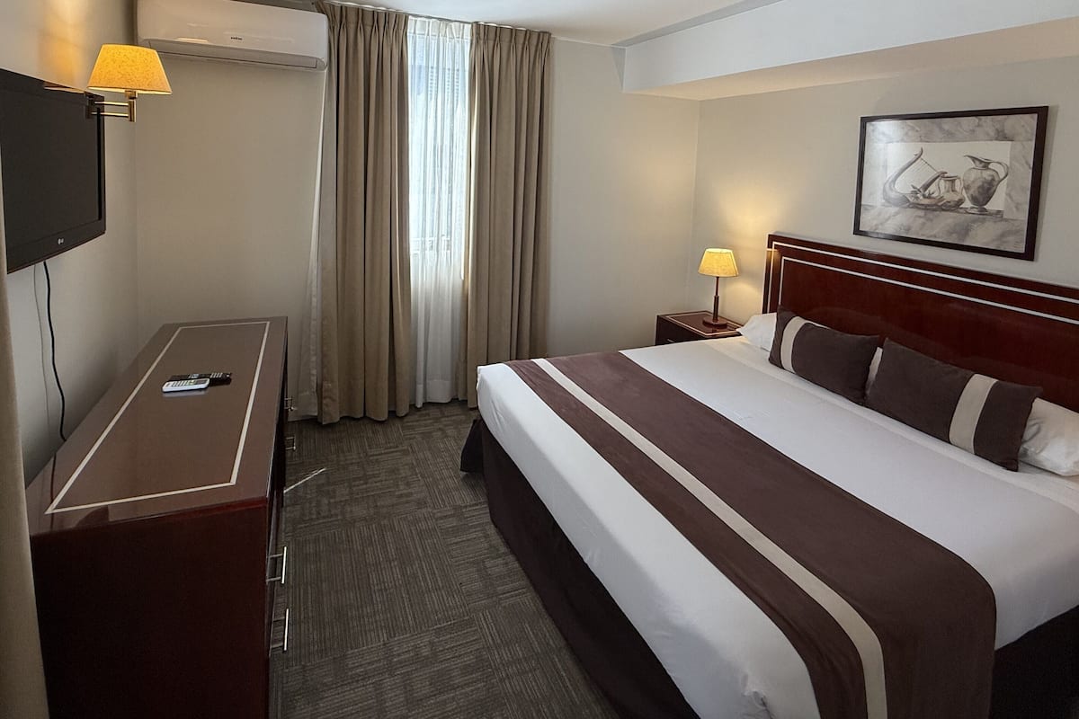 A hotel room with a bed, a desk, a TV, a lamp, and a painting on the wall.
