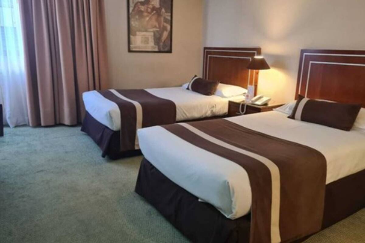 A hotel room with two beds, a nightstand with a lamp, a painting on the wall, and a window with curtains.