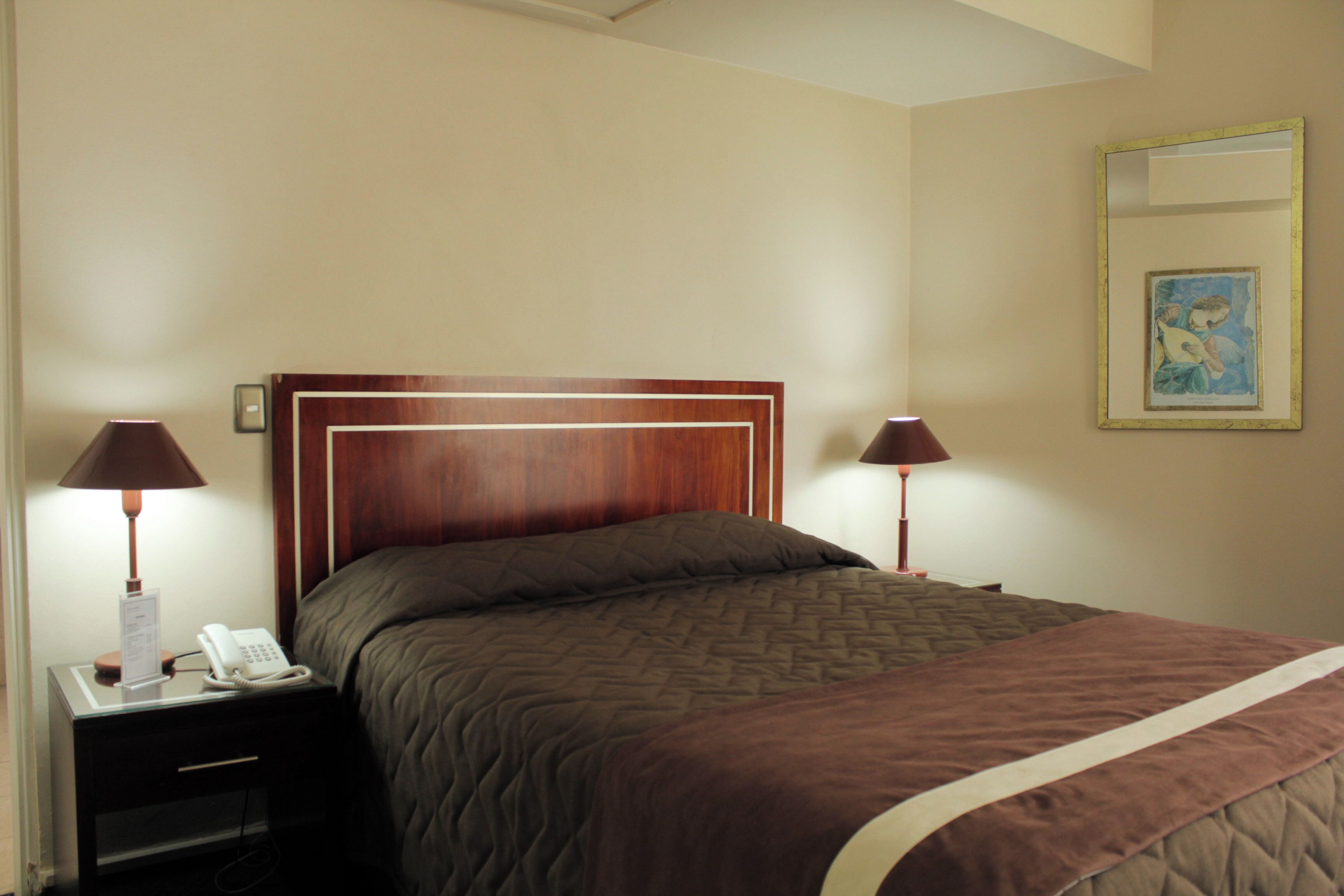 A neatly made bed with a brown quilt and a white accent trim, flanked by two bedside lamps and a nightstand with a phone.