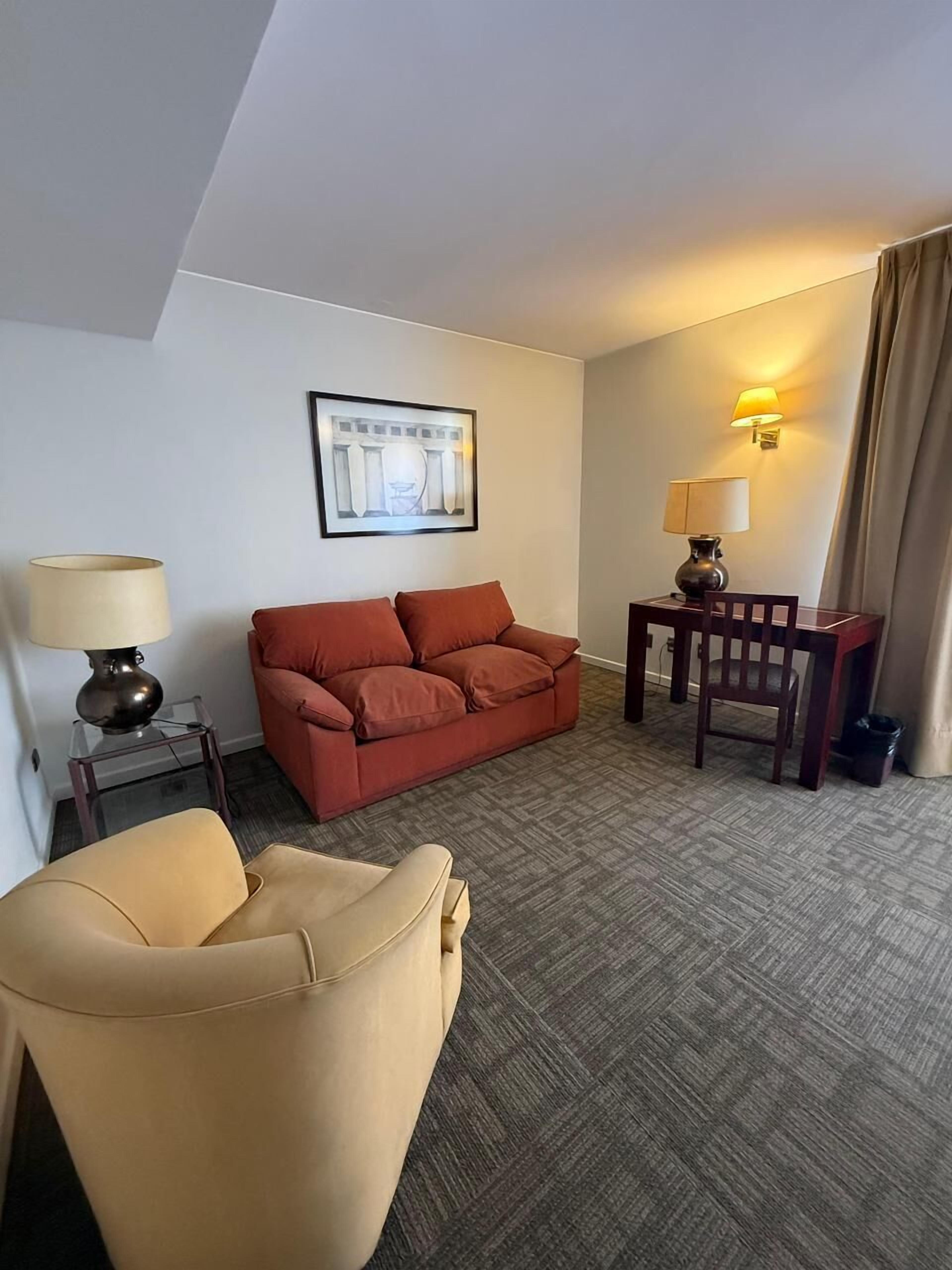 A hotel room with a red sofa, a beige armchair, a small table with a lamp, and a framed picture on the wall.