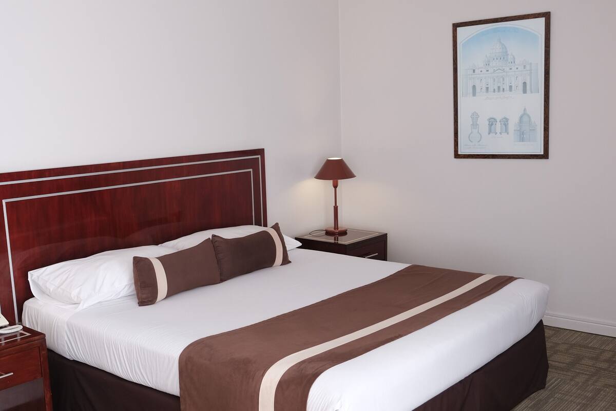 A hotel room with a large bed, a wooden headboard, a nightstand, a lamp, and a framed picture on the wall.