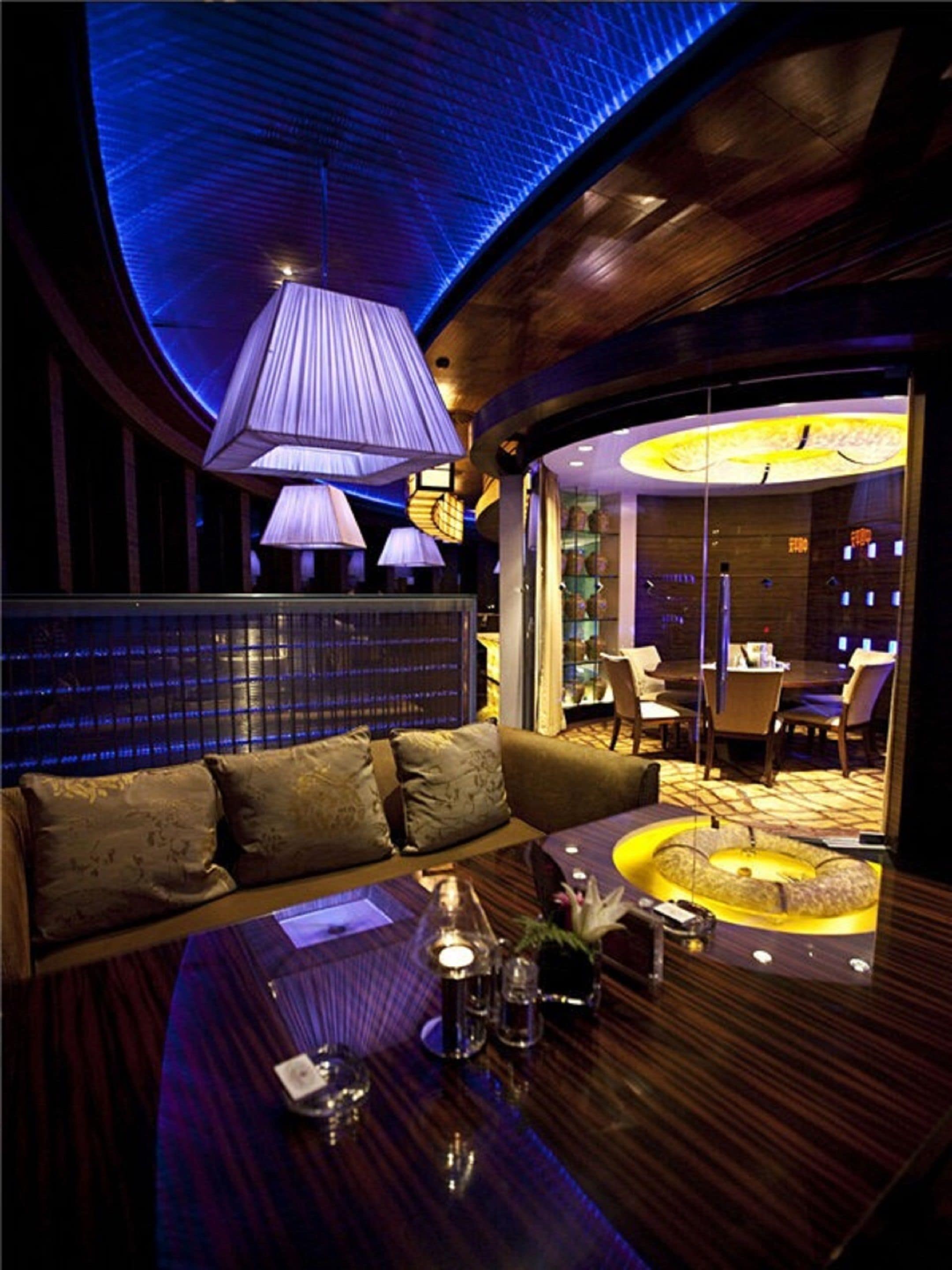 A modern lounge area with a curved wooden bench, a round table with a vase and candles, and ambient lighting.