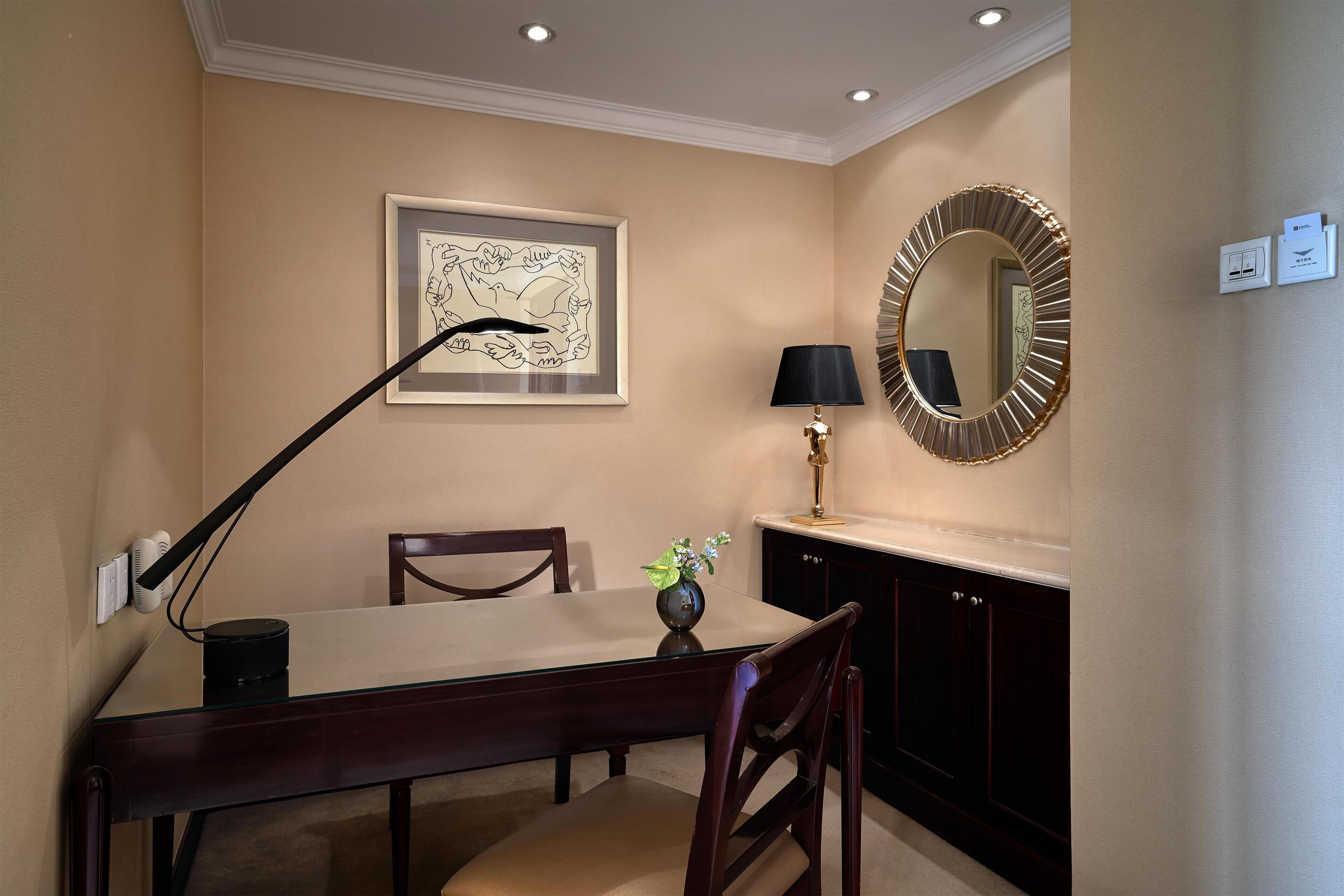 A dining area with a table, chairs, a lamp, a framed artwork, a mirror, and a vase with flowers.