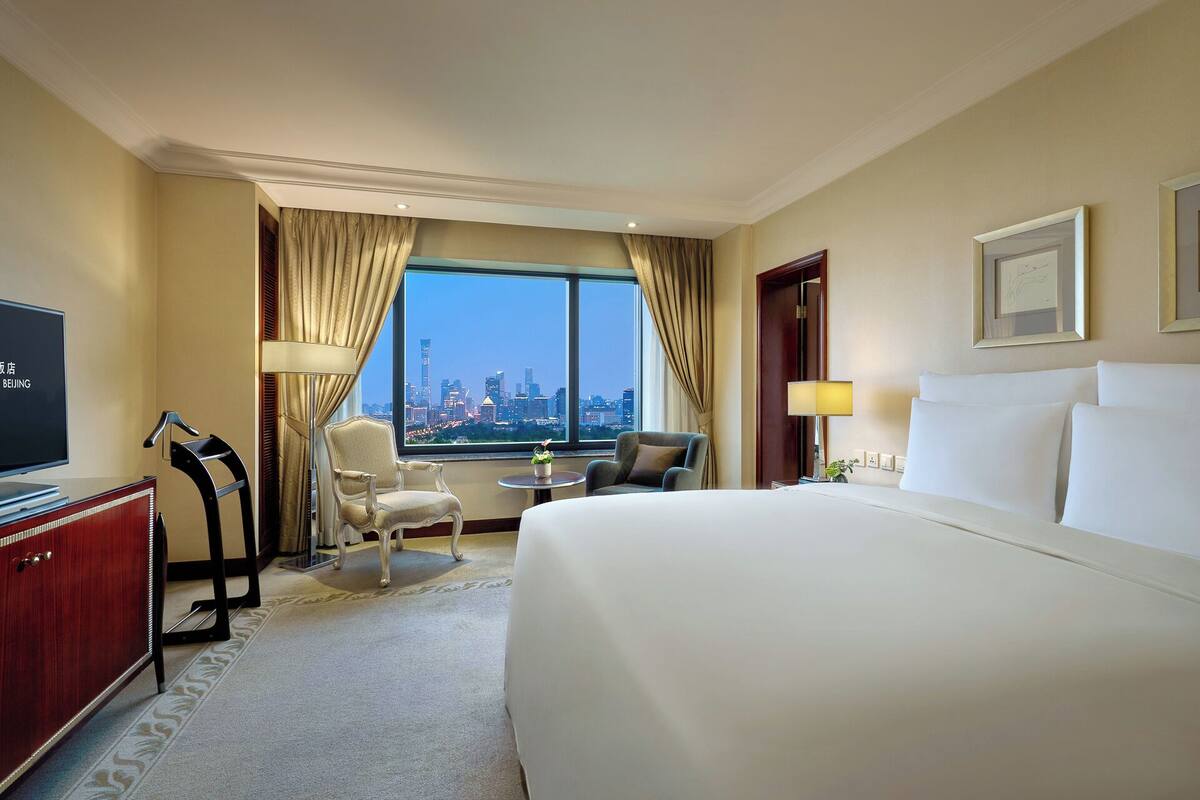 Habitación de hotel con una cama grande, un escritorio con televisor, una silla, una mesita y vistas a un paisaje urbano a través de un ventanal amplio.