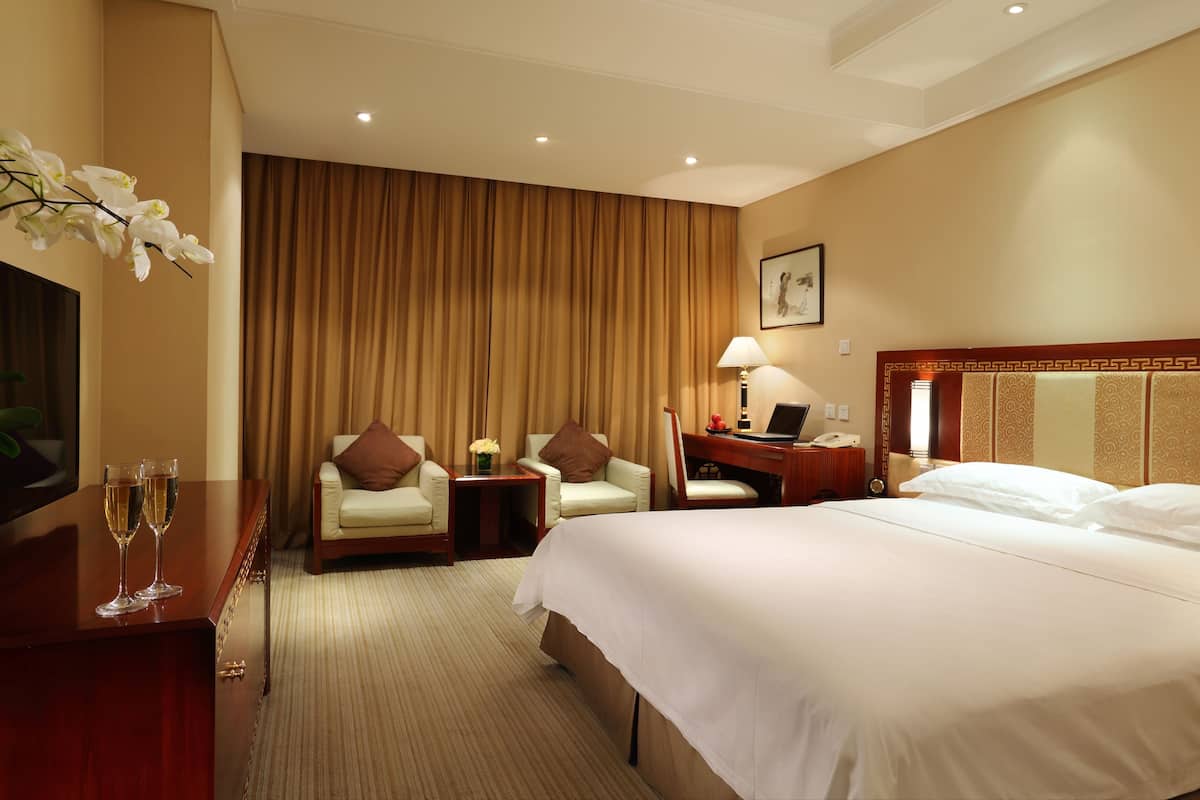 A hotel room with a large bed, a desk with a laptop, two chairs, a small table with a lamp, and a TV on a dresser.