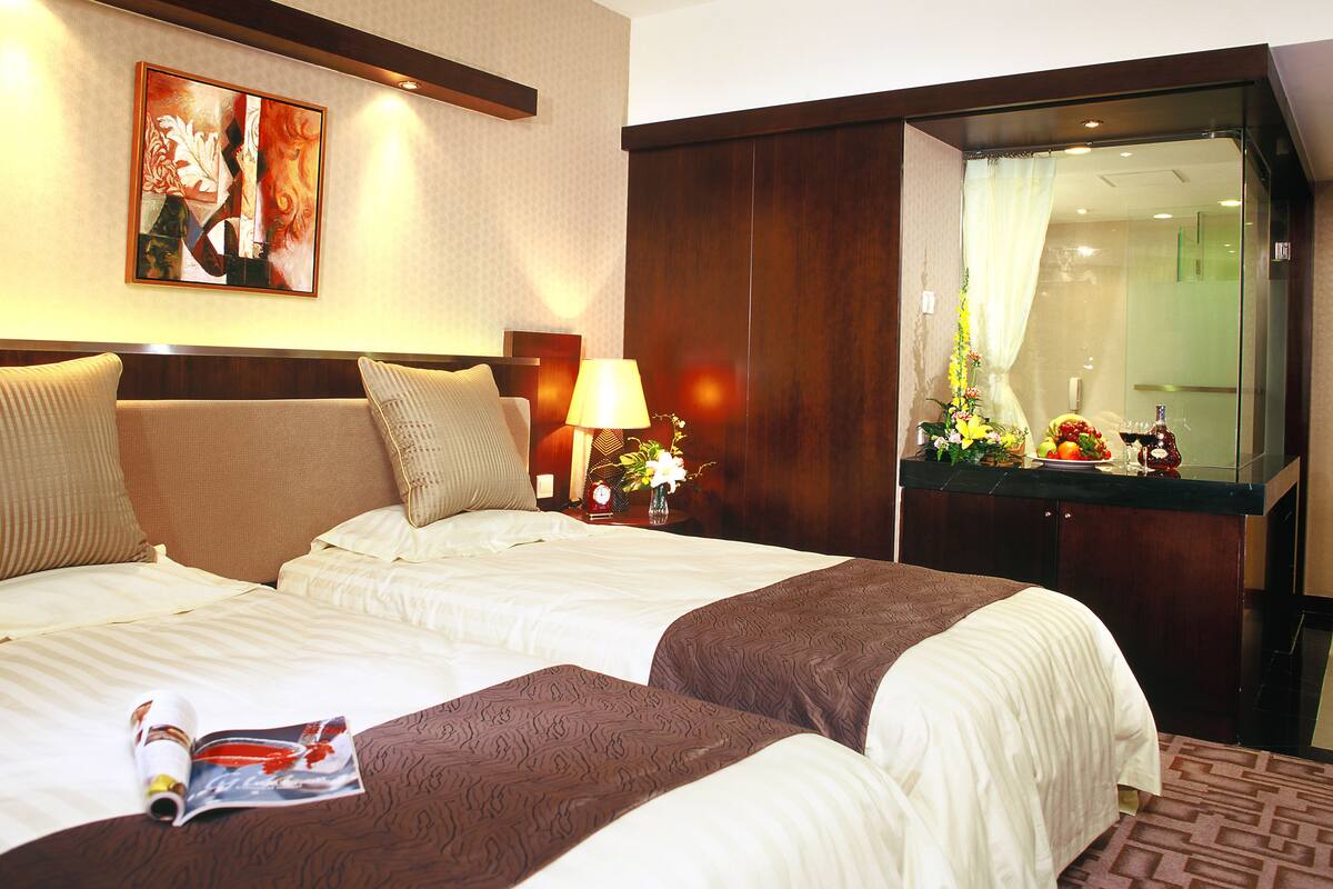 A hotel room with two beds, a nightstand with a lamp, a painting on the wall, and a mirror reflecting the room.