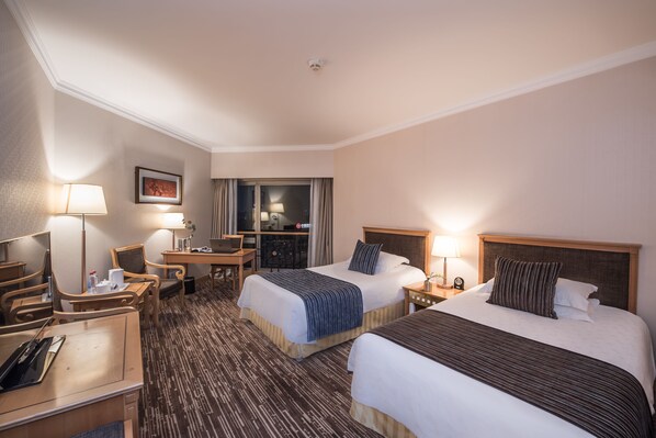 Habitación de hotel con dos camas, un escritorio, una silla, televisión y baño visible a través de una puerta de vidrio.
