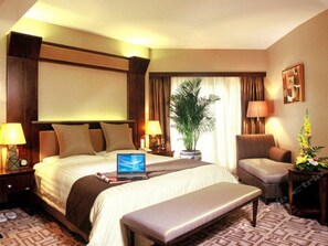 Habitación de hotel con una cama grande, un escritorio con una computadora portátil, una silla, una planta en maceta y una ventana con cortinas.