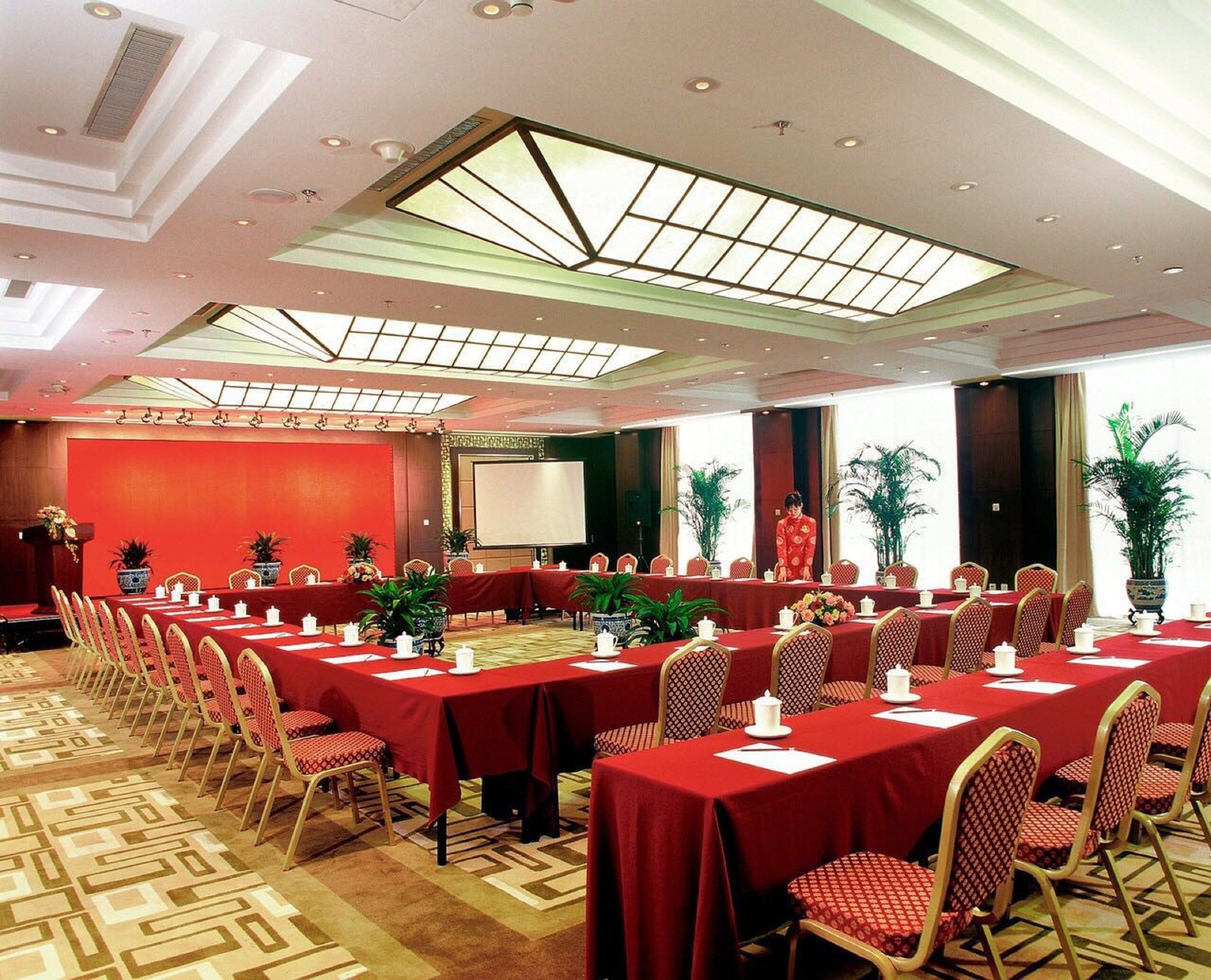 A conference room with multiple tables and chairs, a large screen, and a ceiling with skylights.
