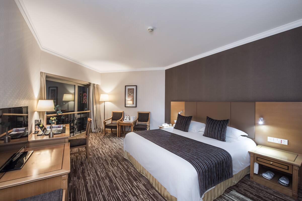 A hotel room with a large bed, a desk with a lamp, two chairs, a small table, and a nightstand with a phone and a lamp.