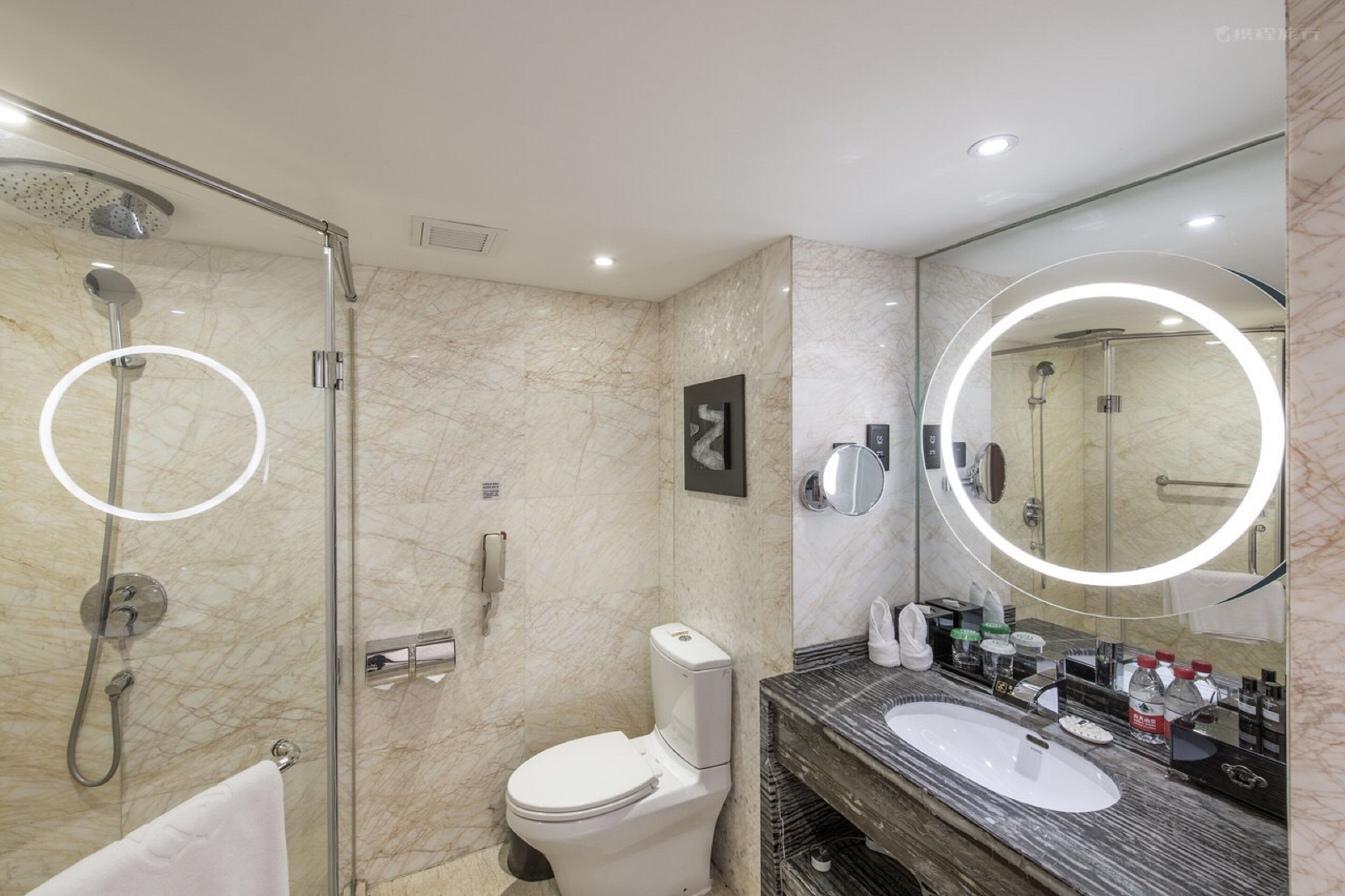 Ein modernes Badezimmer mit einer gläsernen Duschkabine, einer Toilette, einem Waschbecken mit Marmorplatte und einem großen Spiegel mit runder Beleuchtung.