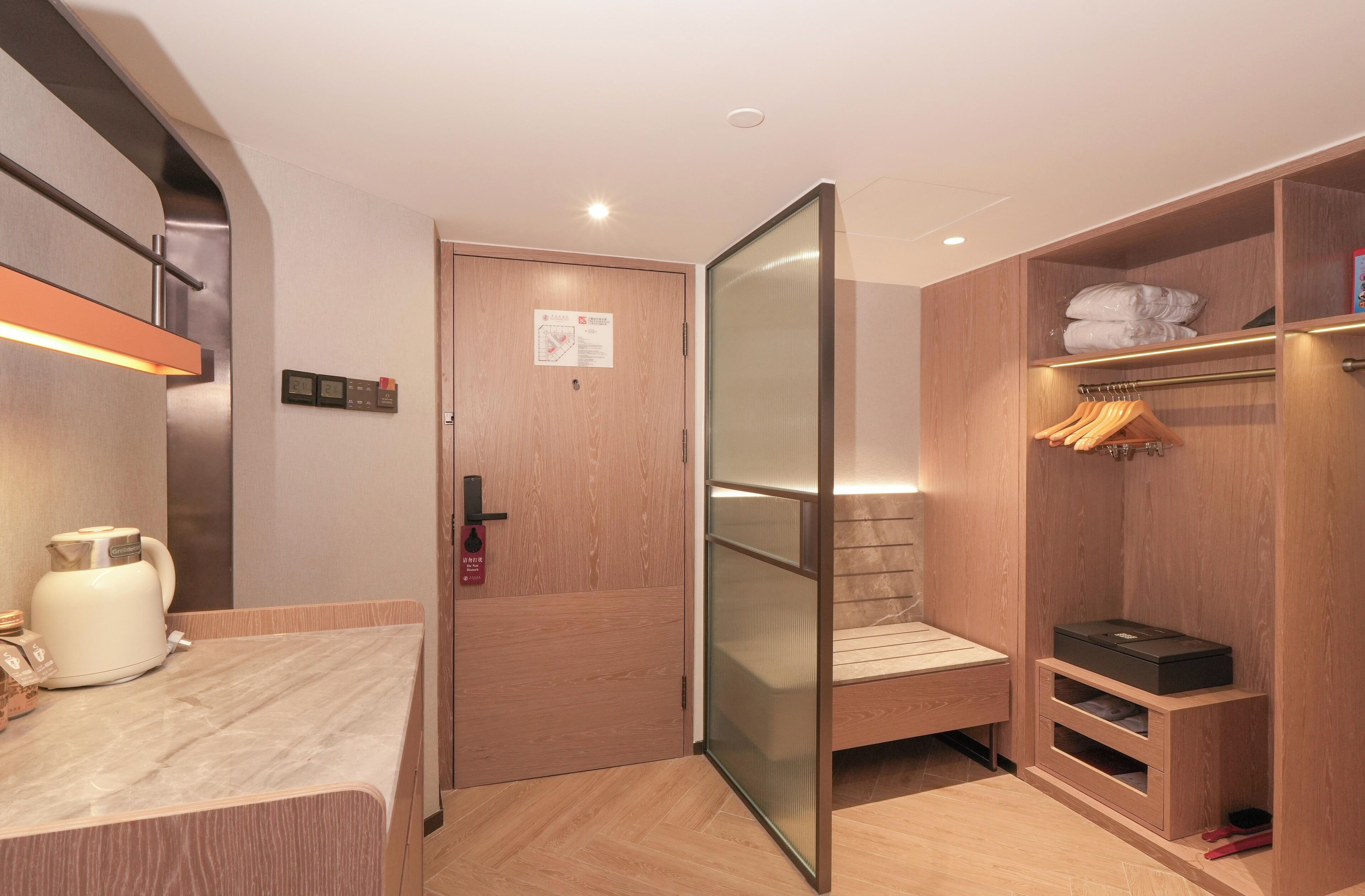 A hotel room with a wooden wardrobe, a bed with a headboard, a small seating area, and a kitchenette with a coffee maker.