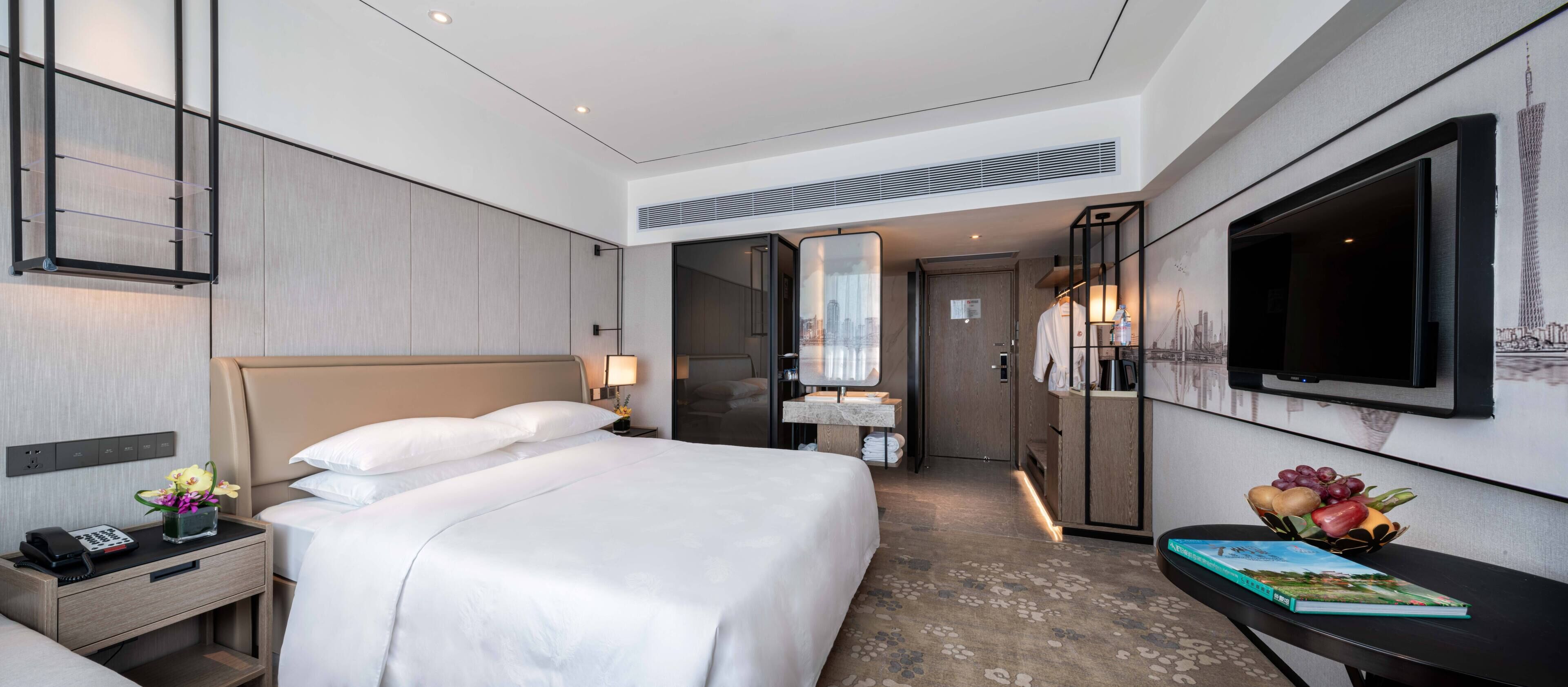 Una habitación de hotel moderna con una cama grande, una mesita de noche con un teléfono, una pequeña mesa con un libro y fruta, un televisor y un espejo.