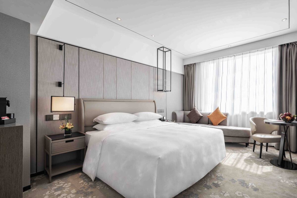 A modern hotel room with a large bed, a desk with a lamp, a small table with fruit, and a TV.