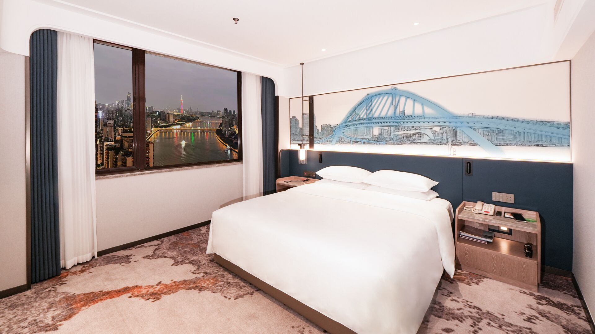 A hotel room with a large bed, a nightstand, a lamp, and a view of a cityscape with a bridge.