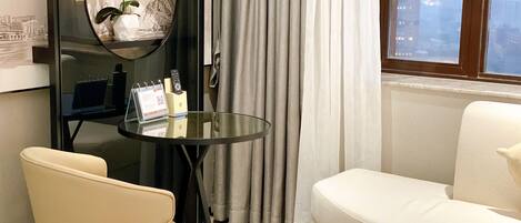 A hotel room with a bed, a round mirror, a small table with a calendar and remote controls, a white sofa, and a window with a city view.