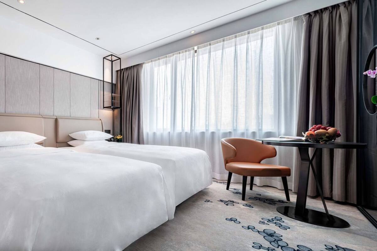 A hotel room with two beds, a desk, a chair, and a large window with curtains.