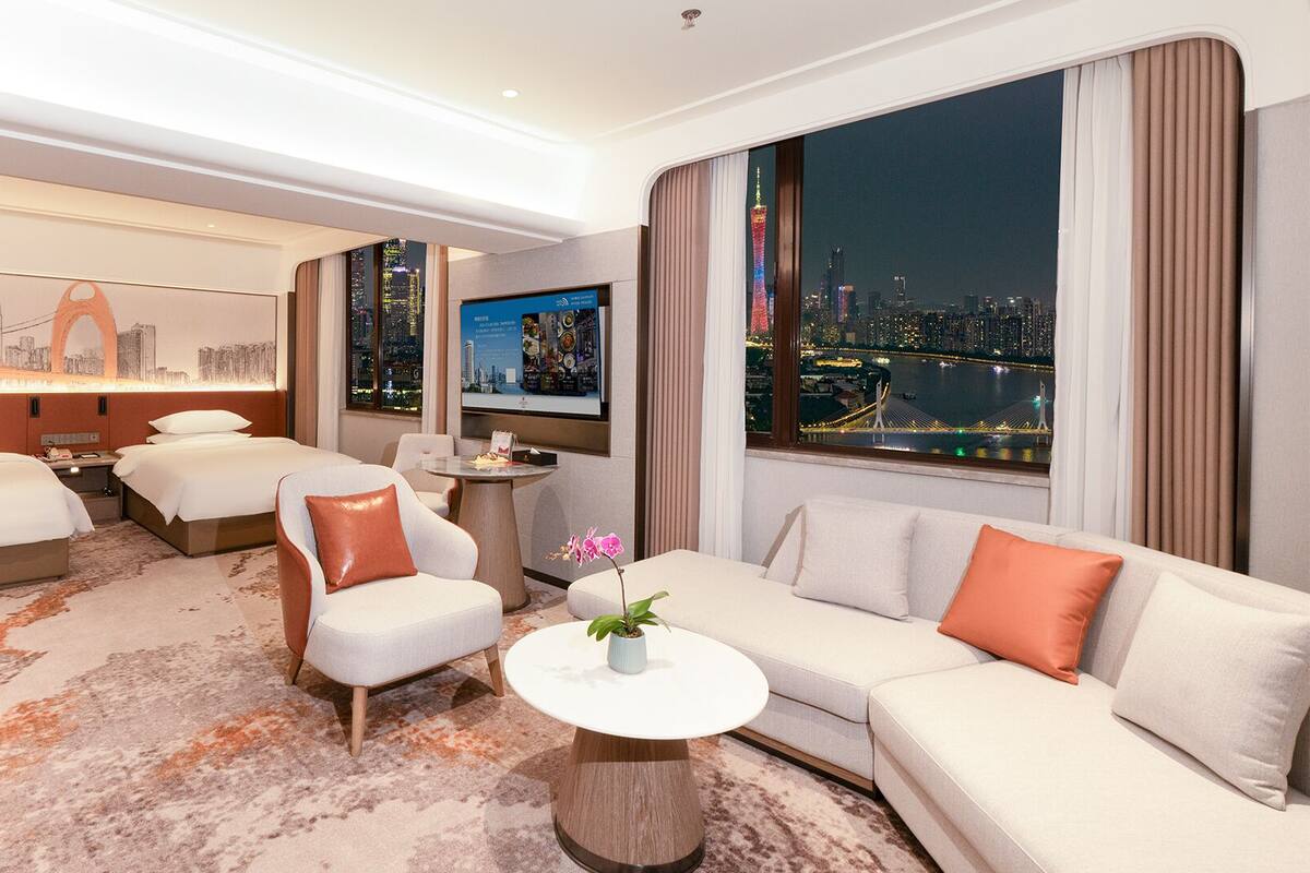 A hotel room with a city view, a bed, a sofa, a chair, a small table, and a TV.