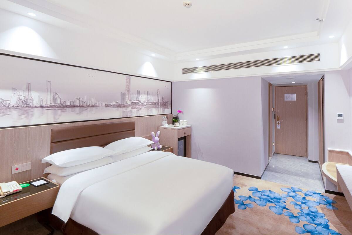 A hotel room with a large bed, a desk with a phone, a small table with a vase, a wall-mounted TV, and a cityscape artwork.