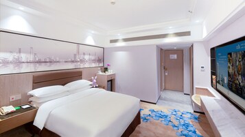 A hotel room with a large bed, a desk with a phone, a small table with a vase, a wall-mounted TV, and a cityscape artwork.