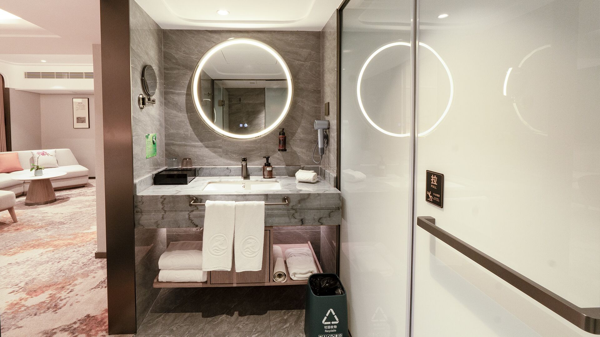 A modern bathroom with a marble vanity, a round mirror with lighting, a towel rack, and a trash bin.