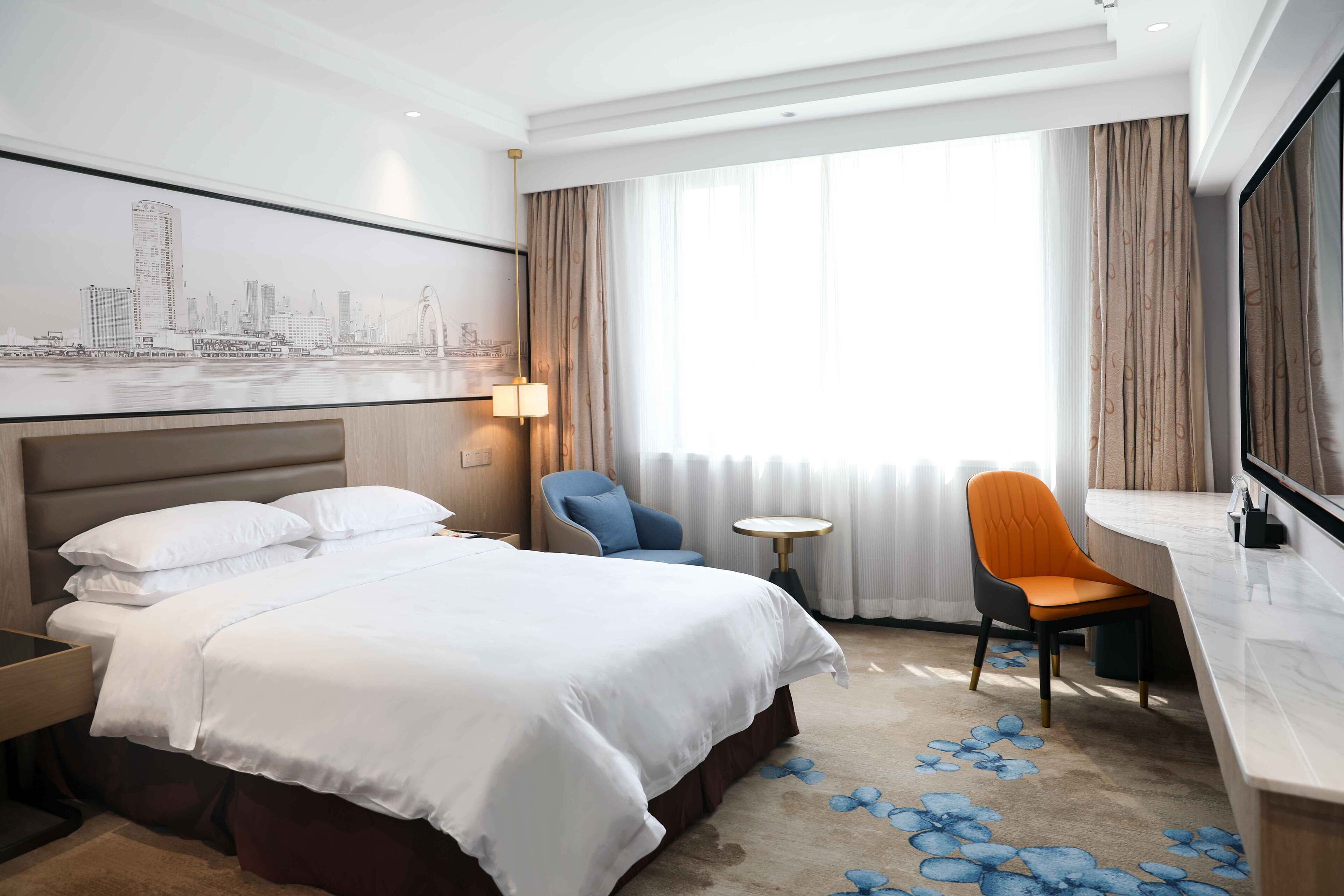 A hotel room with a large bed, a desk, a chair, a small table, and a cityscape view.