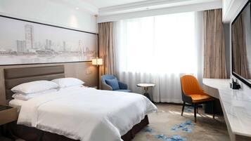 A hotel room with a large bed, a desk, a chair, a small table, and a cityscape view.