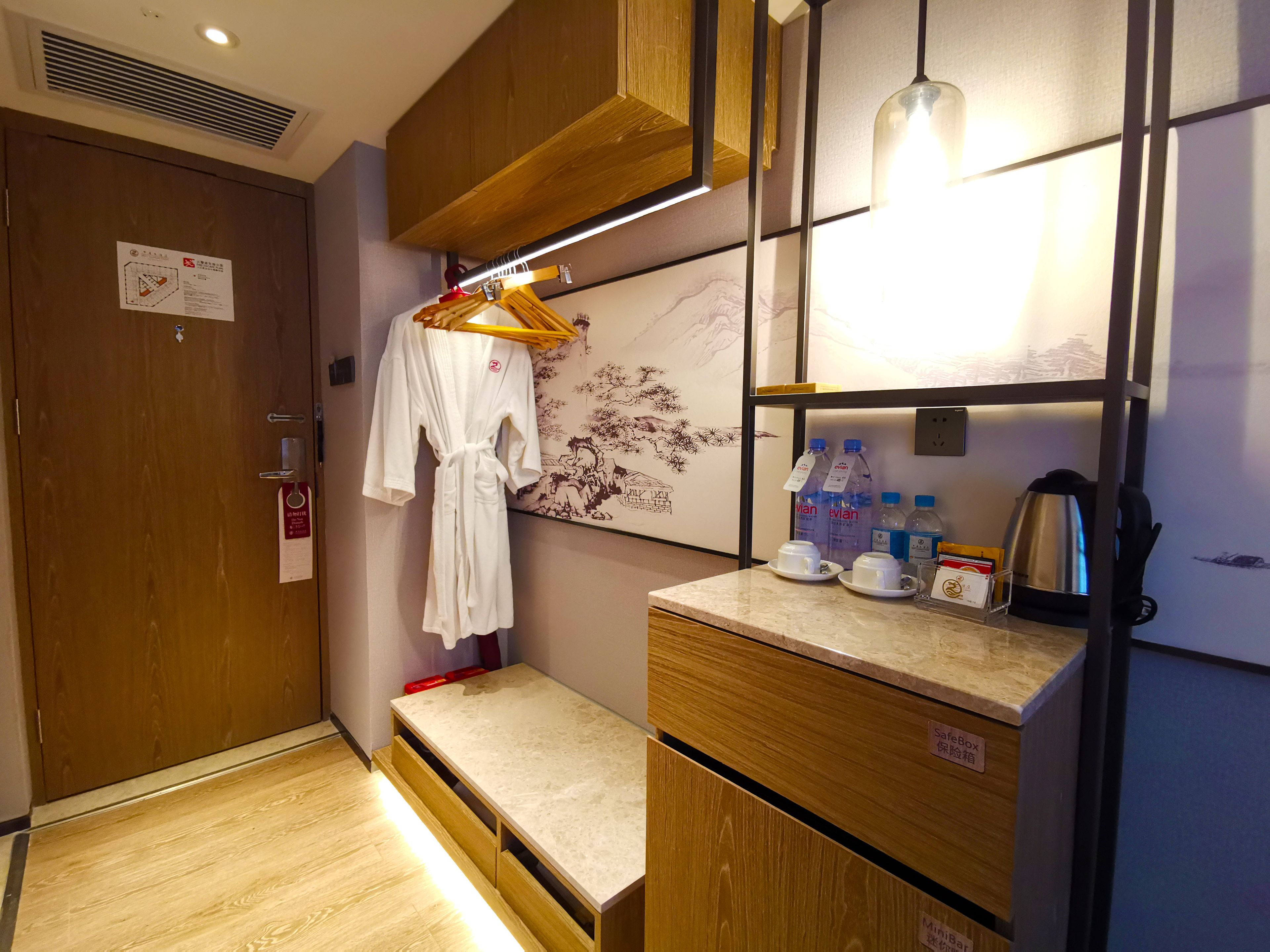 A hotel room with a wooden wardrobe, a bench, a robe hanging on a rack, a shelf with water bottles and a kettle, and a wall with a decorative mural.