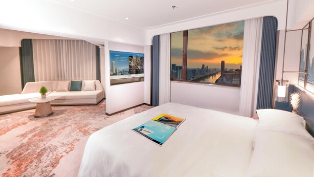 Una camera d'hotel moderna con un letto grande, un divano, un tavolino con una pianta, un quadro con una veduta cittadina e vista su un tramonto.