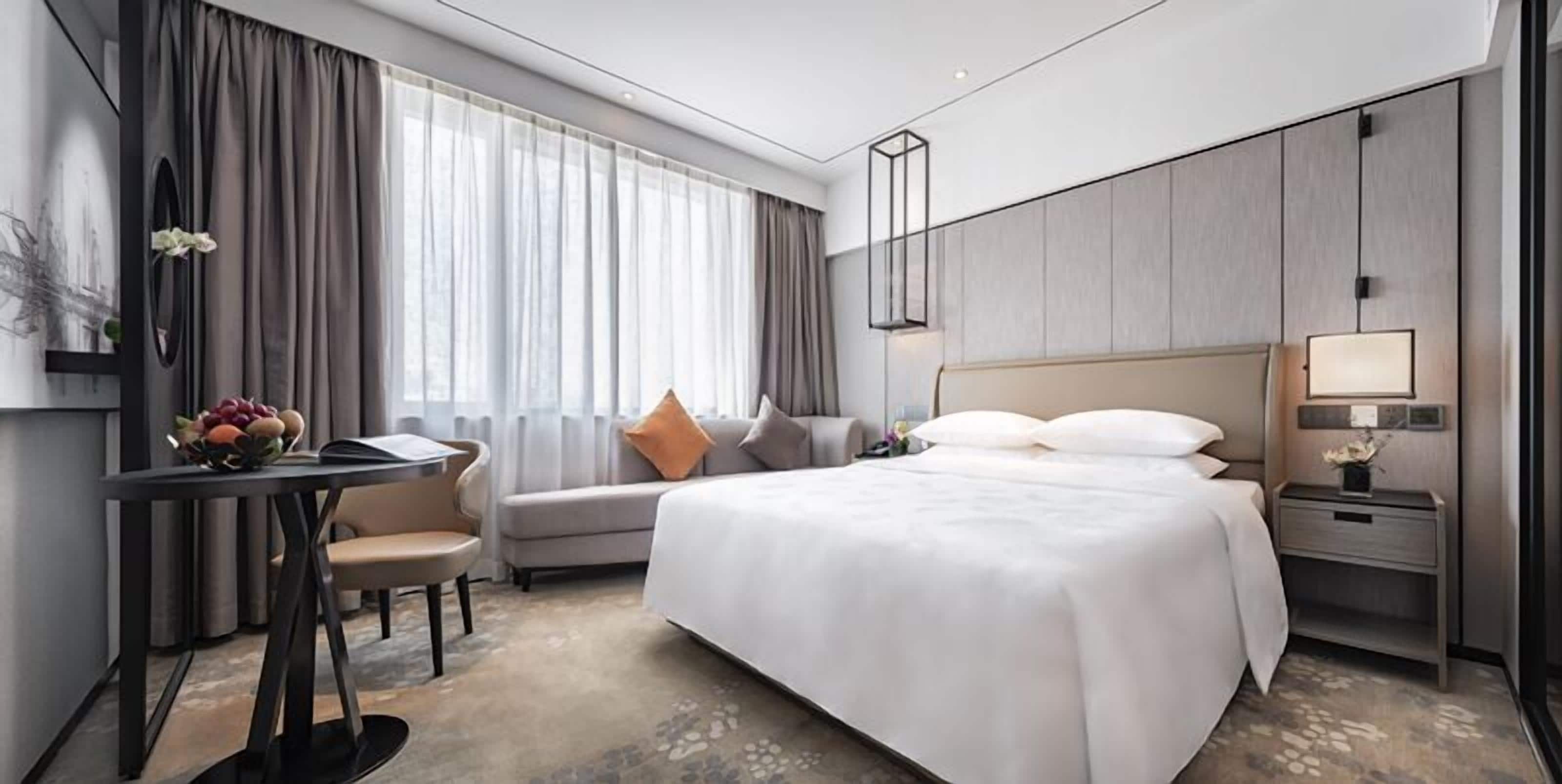 Una habitación de hotel moderna con una cama grande, un escritorio con silla, un sofá y una mesita de noche con lámpara.