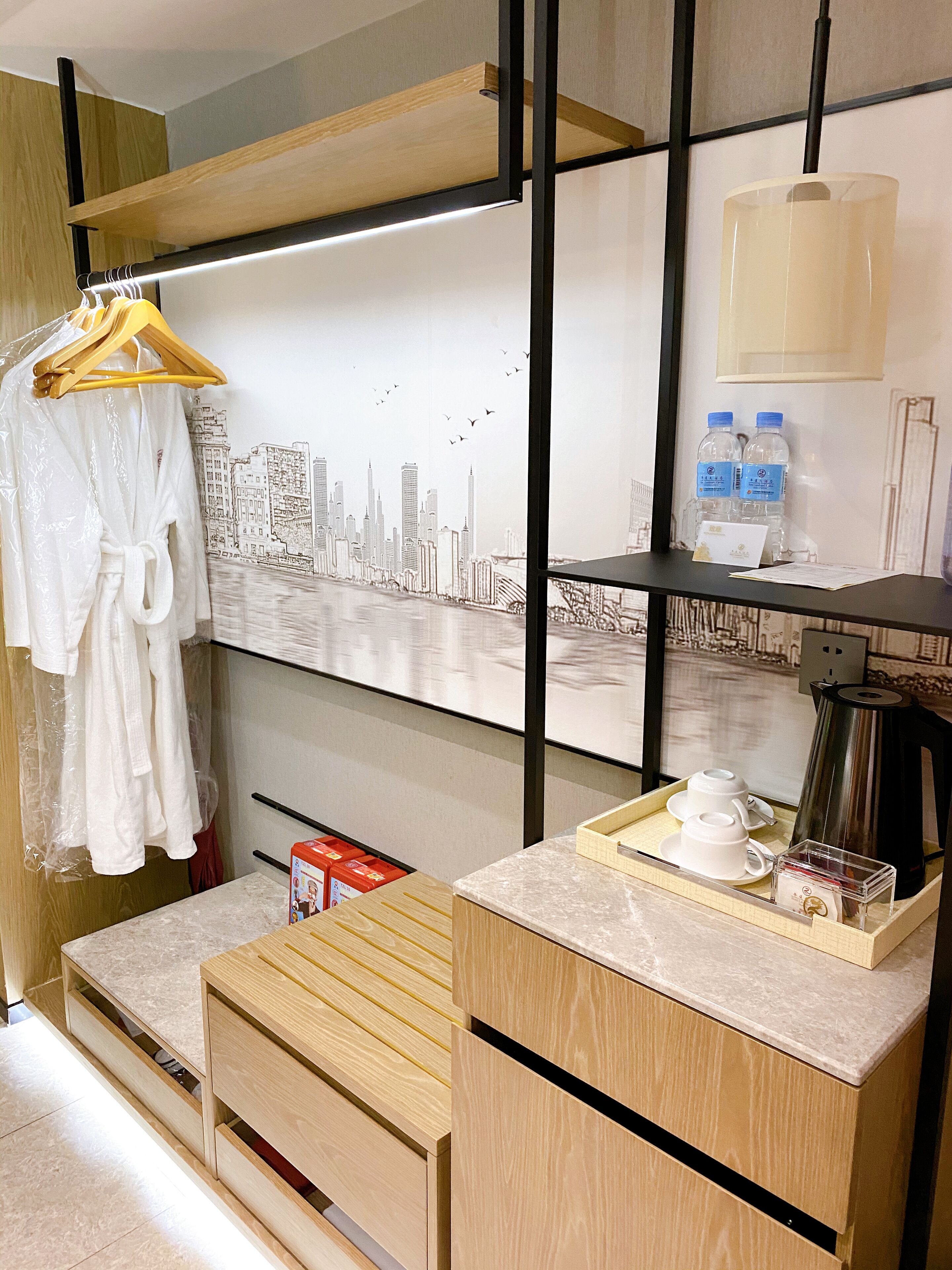 A hotel room with a wooden desk, a painting of a city skyline, a hanging robe, and a coffee station.