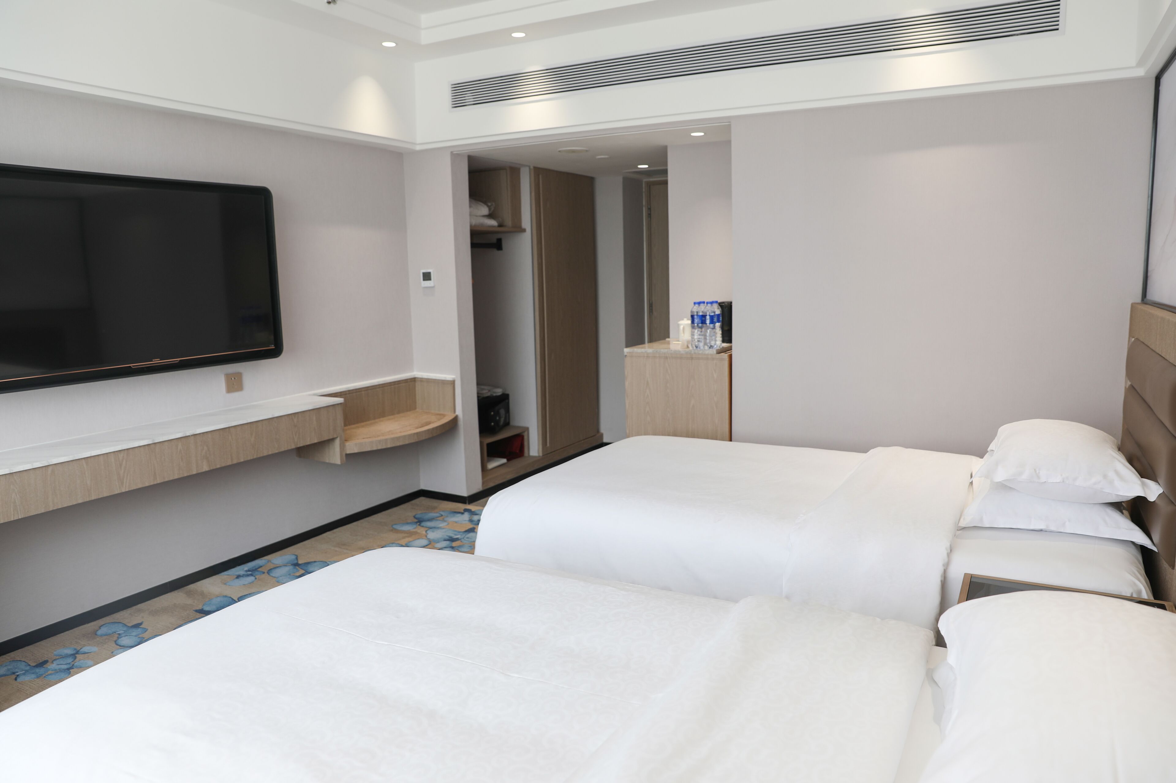 A hotel room with two beds, a TV mounted on the wall, a small shelf with toiletries, and a mirror above the bed.