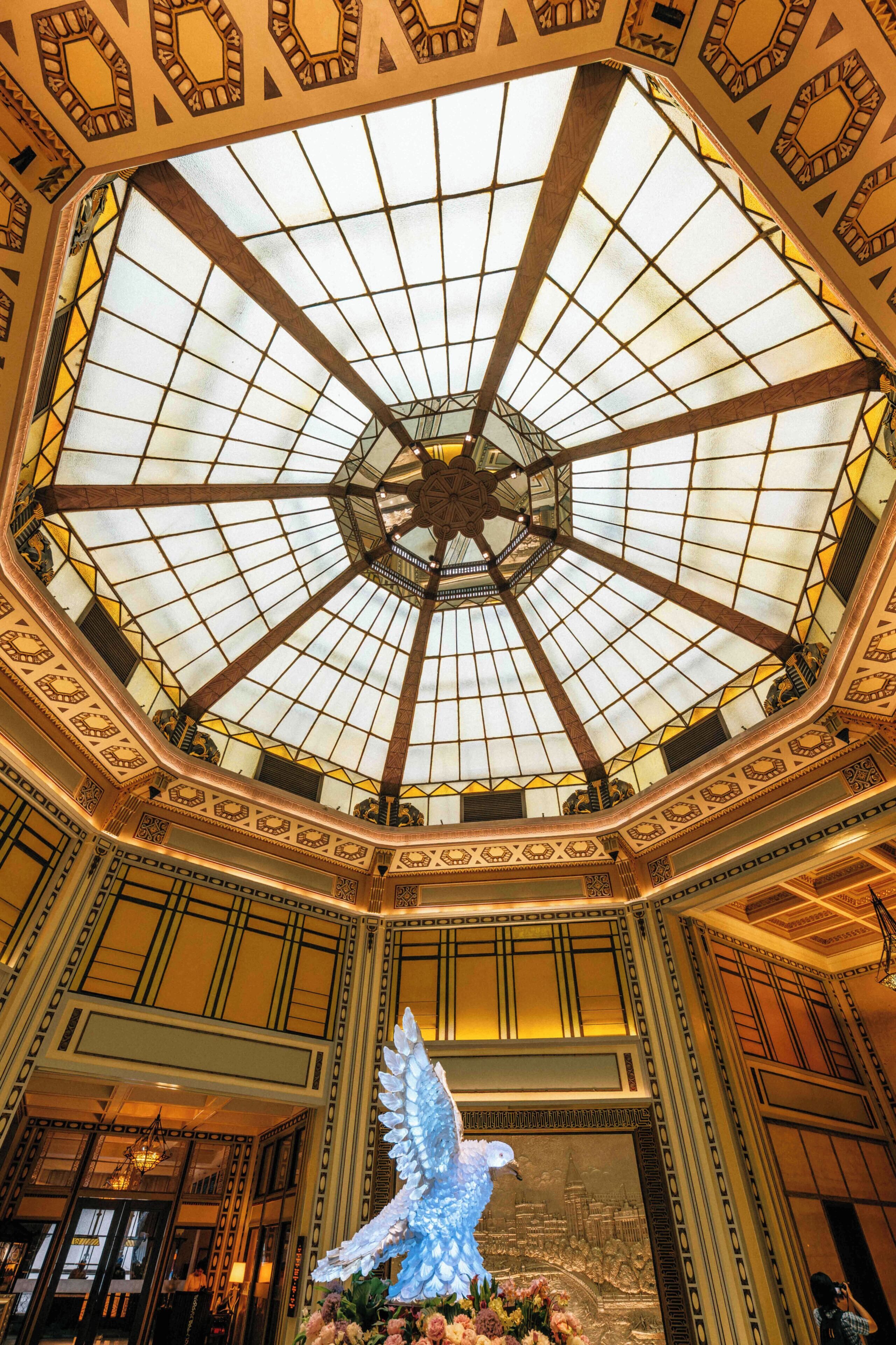 A grand hall with a large glass dome ceiling, ornate wall designs, and a central sculpture of a dove.