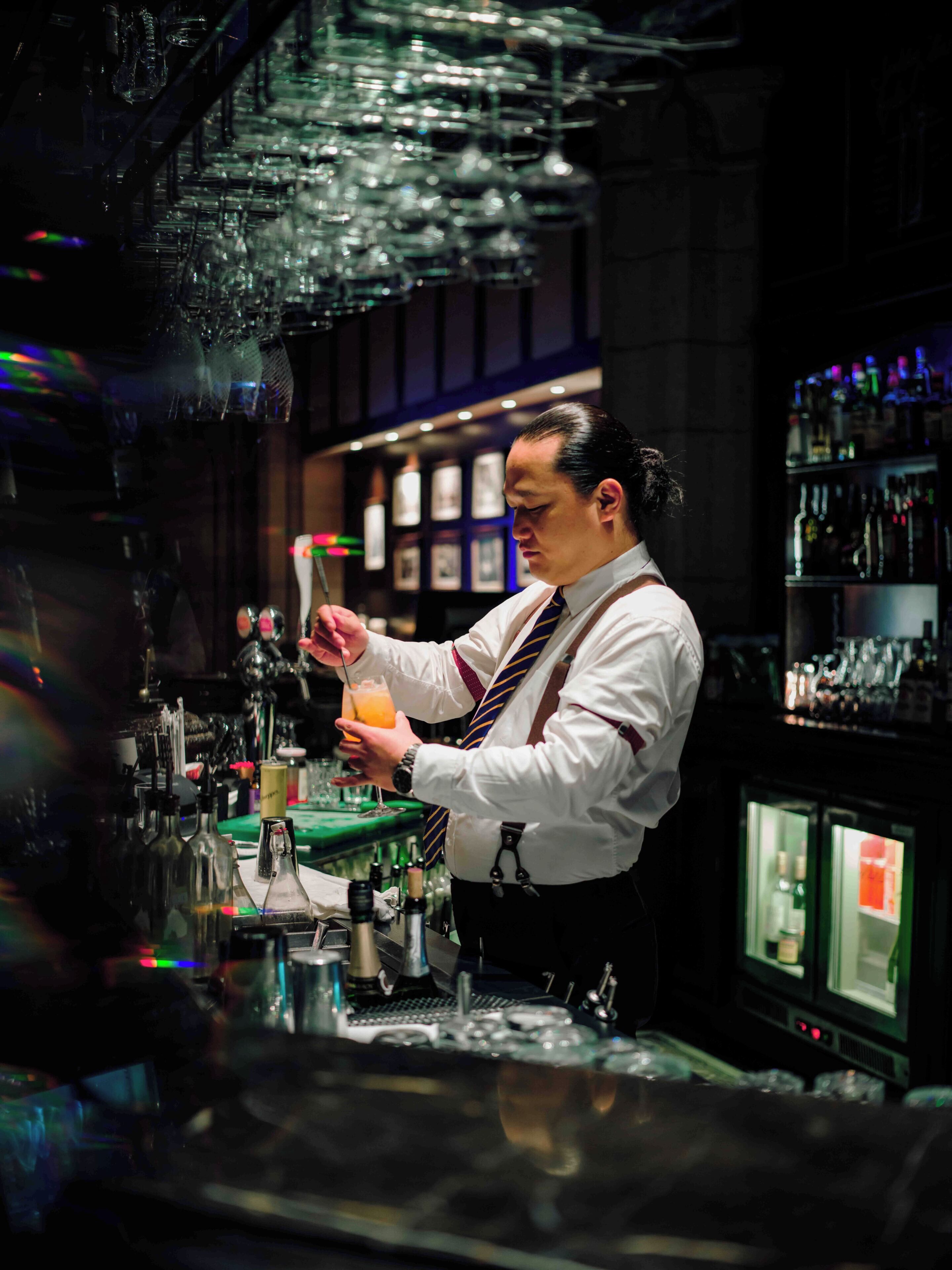 A bartender at a bar with a well-stocked shelf of bottles and a refrigerated display.