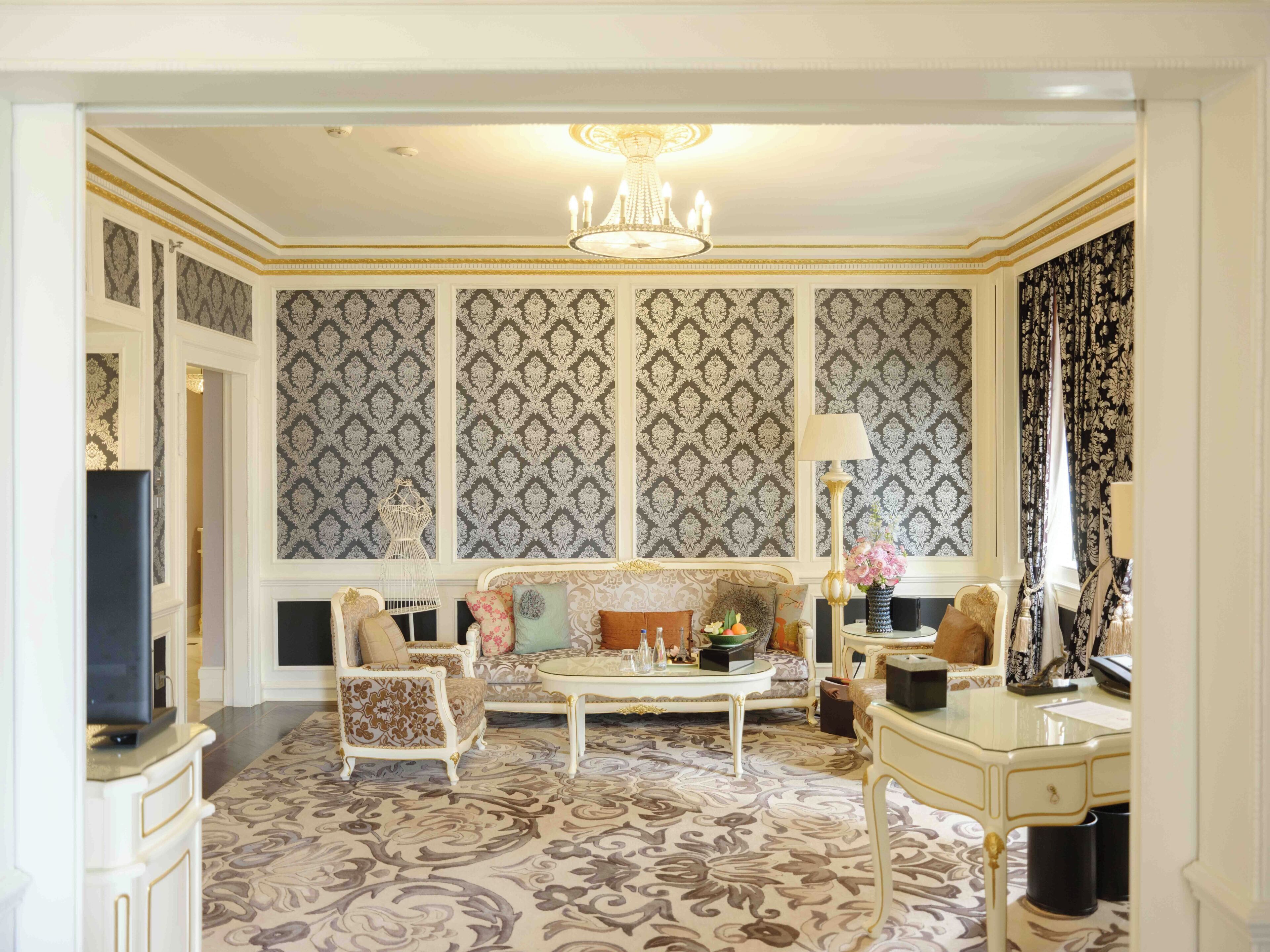 Une pièce avec un papier peint à motifs, un lustre, un canapé, une table basse et un bureau.