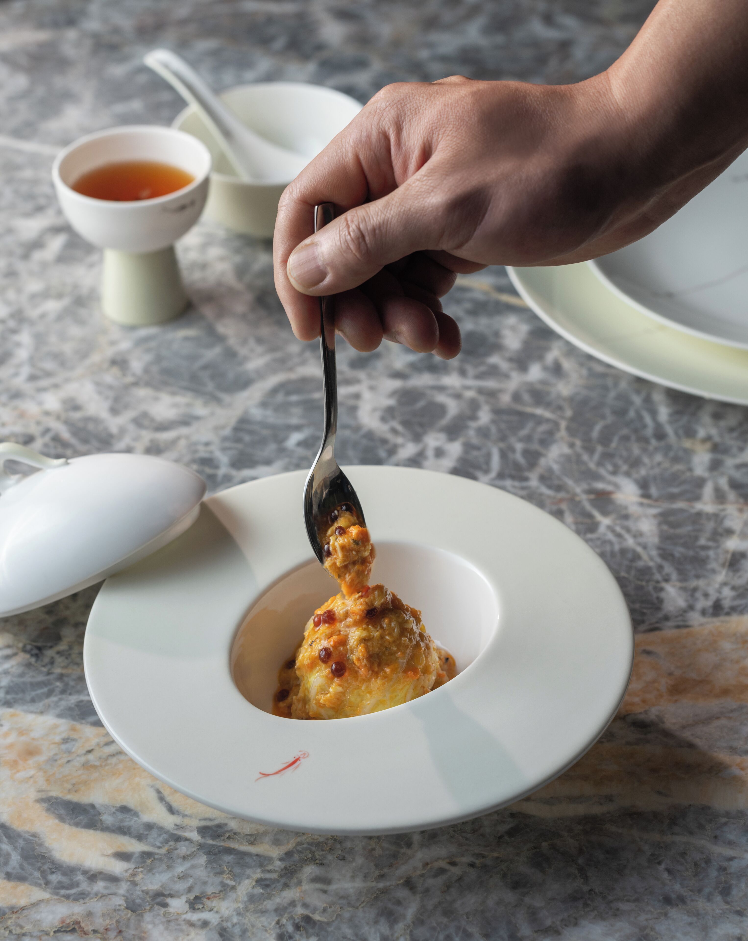 A hand holding a spoon over a white bowl containing a food item, with a small bowl of sauce and a spoon in the background on a marble surface.