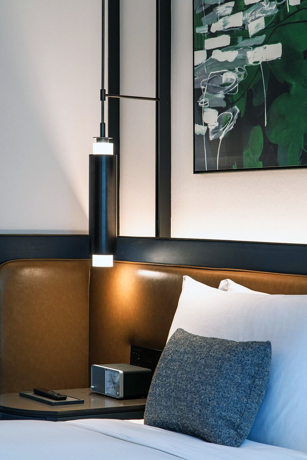 Ett modernt hotellrum med en brun läderklädd sänggavel, ett nattduksbord med en klocka, en fjärrkontroll och en inramad abstrakt målning på väggen.