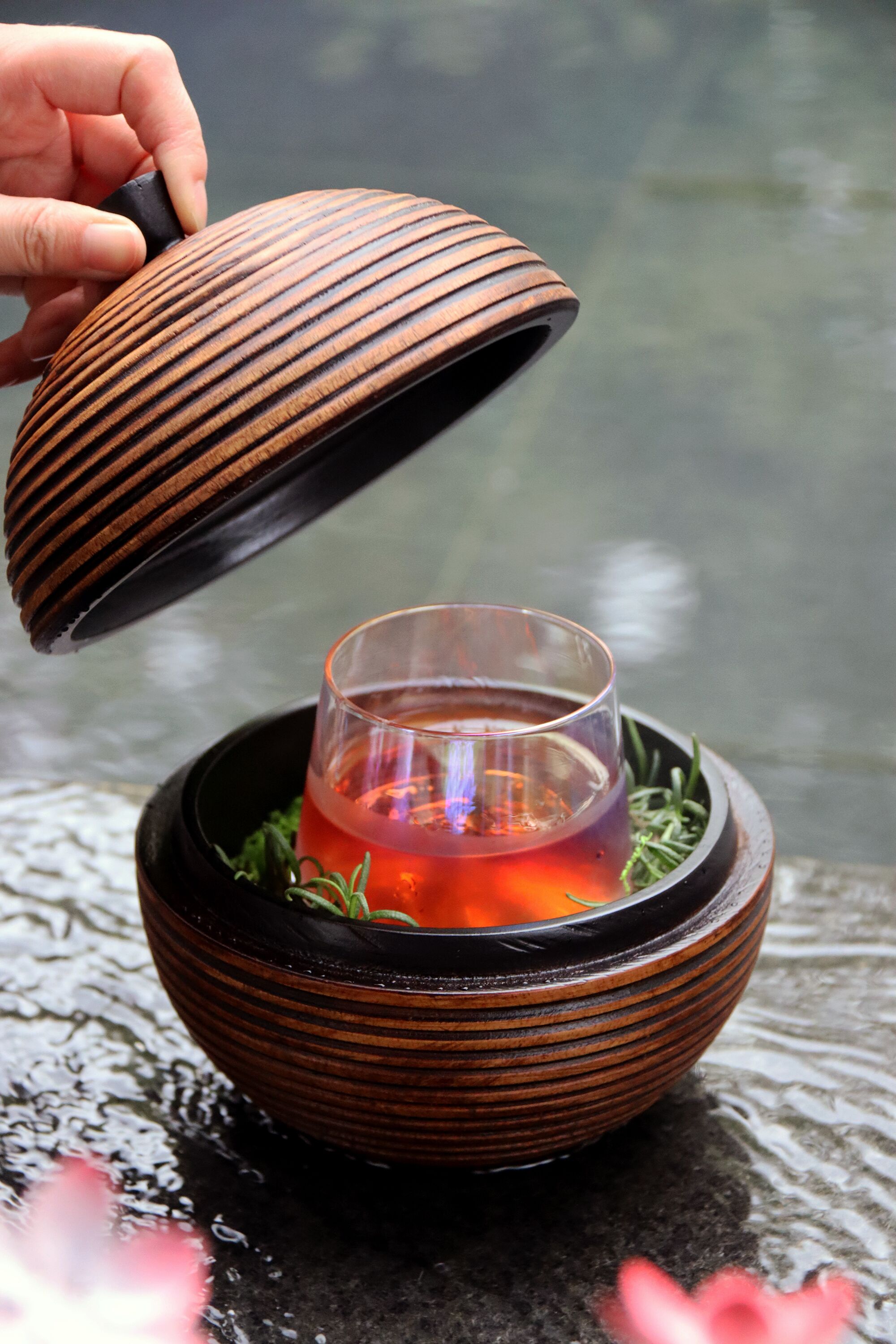A hand lifting a lid from a ceramic bowl containing a glass of tea with herbs.