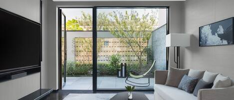 A modern living room with a flat-screen TV, a sofa, a coffee table, and a large sliding glass door leading to a balcony with a view of greenery and a fence.