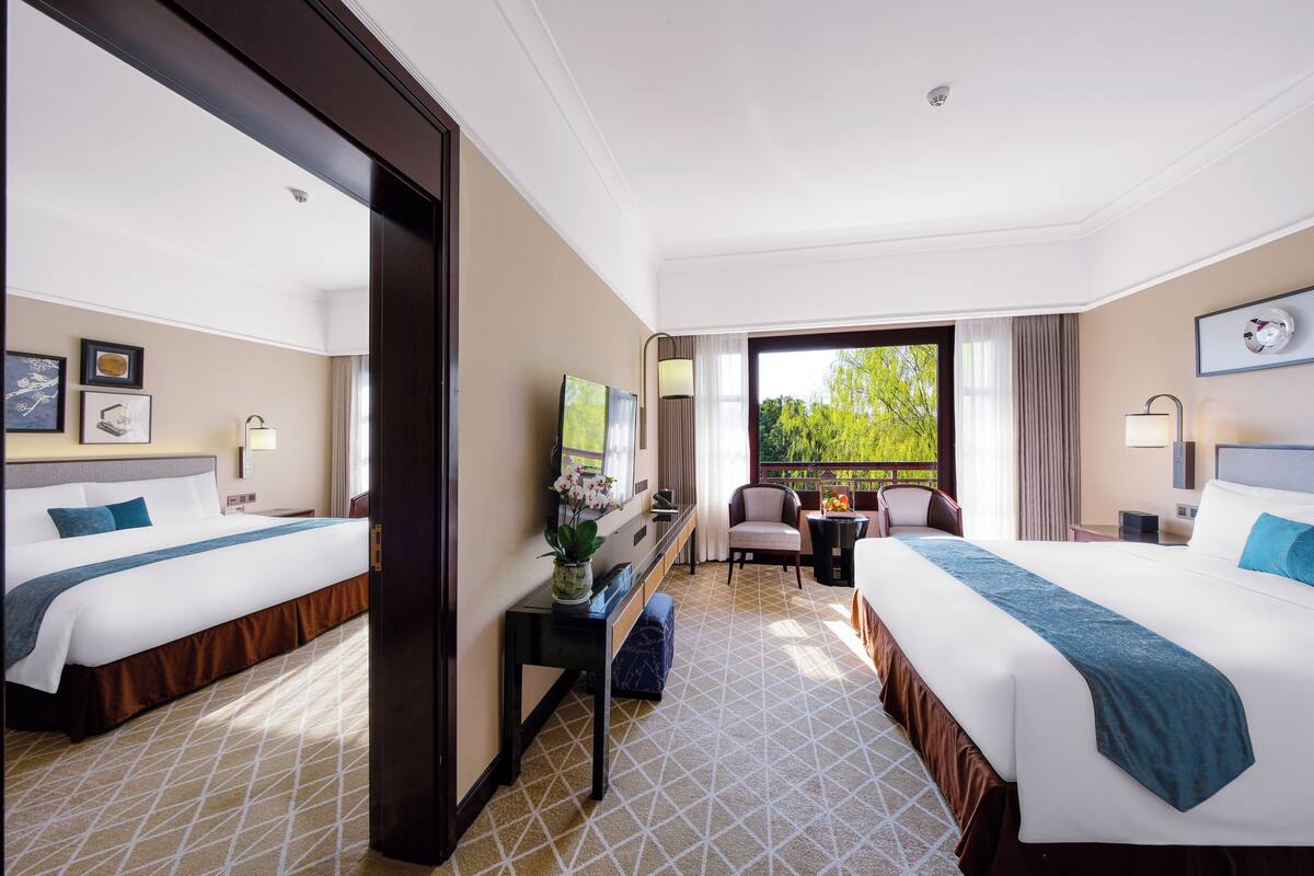 A hotel room with two beds, a desk with a chair, a small table, and a view of greenery outside.