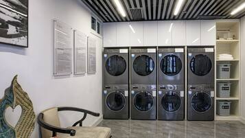 A modern laundry facility with four front-loading washing machines, a seating area with a chair and a table, and a shelf with folded towels.