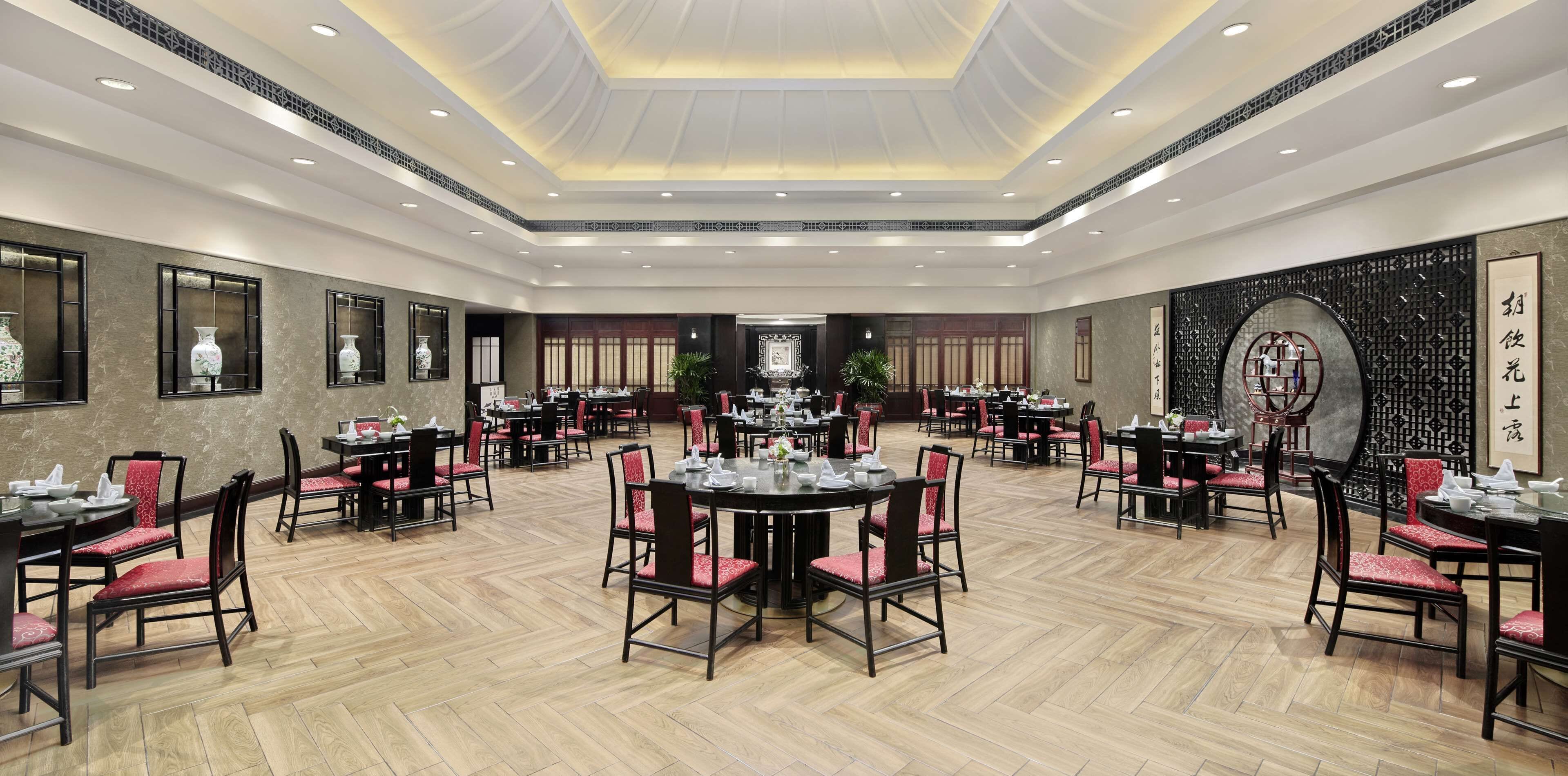 A spacious dining area with multiple round tables and chairs, a high ceiling with recessed lighting, and traditional decor elements.